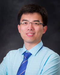 A man smiles in to a camera. He is wearing pale blue official shirt and a checked deep blue tie.